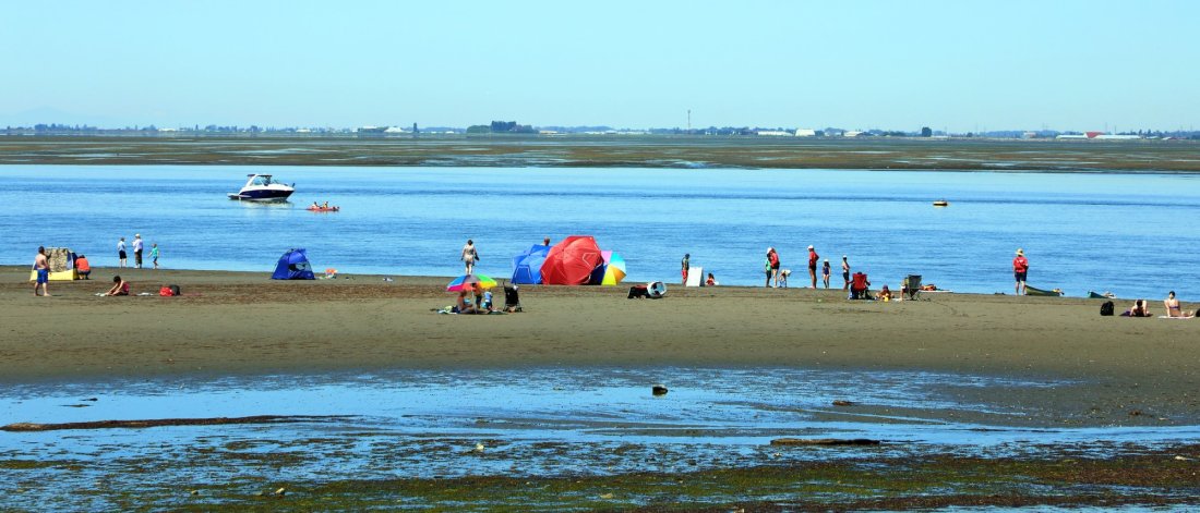 Families and individuals enjoy a sunny day at Crescent Beach, with a boat cruising nearby and a colorful beach umbrella set up on the shore.