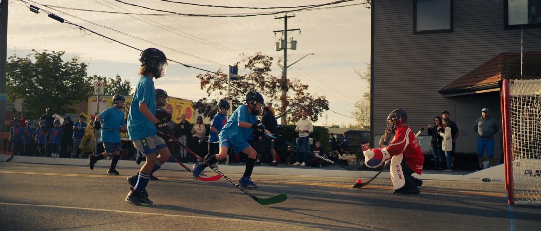 Children playing street hockey during a community event in Cloverdale, with spectators watching from the sidelines as a goalie in red gear makes a save.