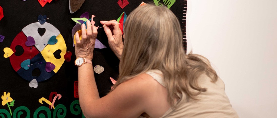 A crouching woman attaches a piece of felt to a blanket hanging on the wall.