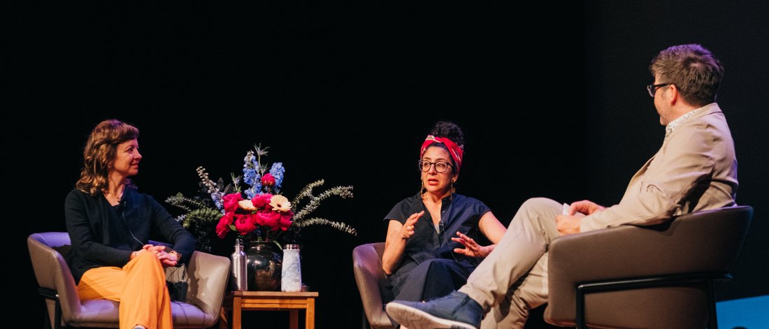 Artists Jaimie Robson and Maya Ersan sit on chairs on a stage, talking about their exhibition. Curator Jordan Strom sits across from them as moderator. A vase of flowers is placed on a table between the two artists.