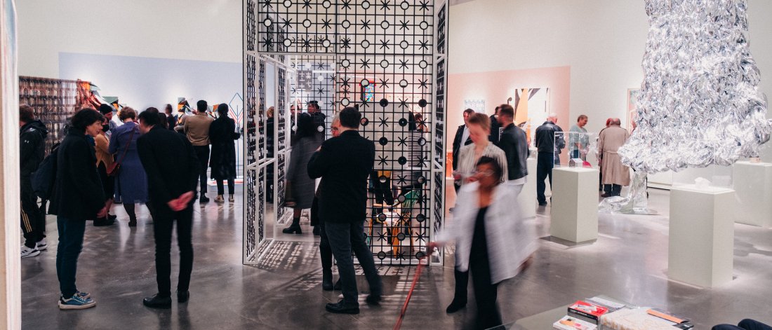 A group of visitors at an opening reception in Surrey Art Gallery. There are many sculptural and architectural pieces in the middle of the space, as well as a large aluminum-looking sculpture hanging from a wall.