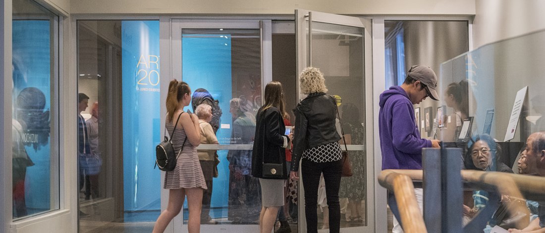 A group of visitors mingle at the entrance to Surrey Art Gallery. The door is open and more visitors are inside the exhibition space where a wall is painted bright blue.