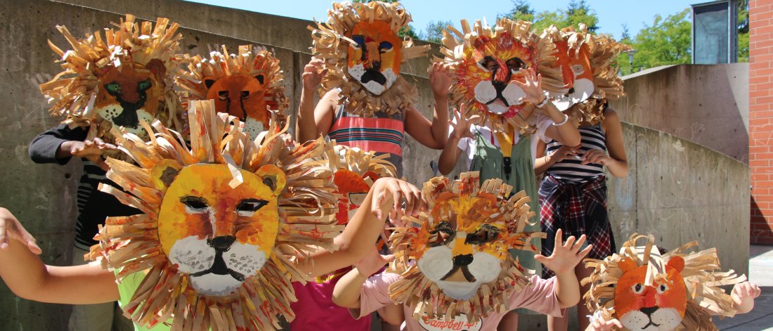 A group of kids stand and kneel in front of a concrete wall with handmade lion masks on their faces and arms out in claw pose.