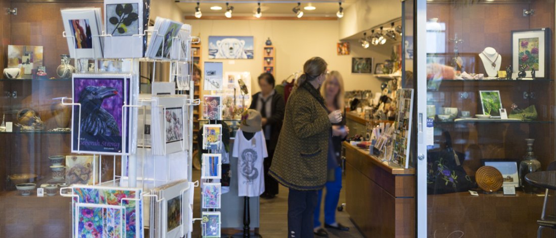 Looking into the Surrey Art Gallery Association's gift shop where a few shoppers are seen mingling and browsing through art racks and displays. A turnstile of art cards is featured on the left side, outside the open doors.