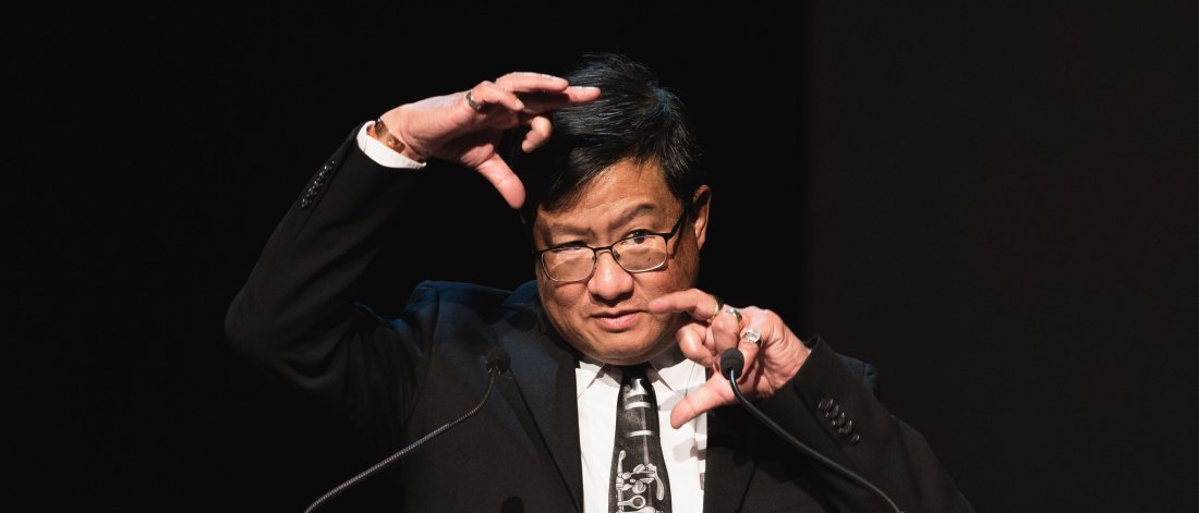 Walter Quan stands behind a podium in a suit and tie making a face and doing expressive hand gestures