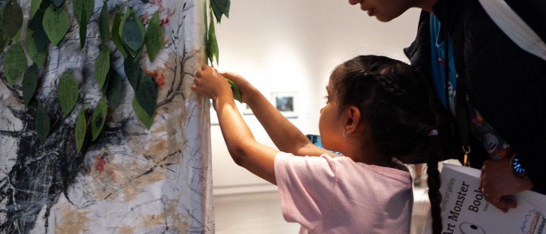 A child pins a leaf onto a canvas with a textured tree image in a gallery space while a caregiver stands behind, leaning in to watch.