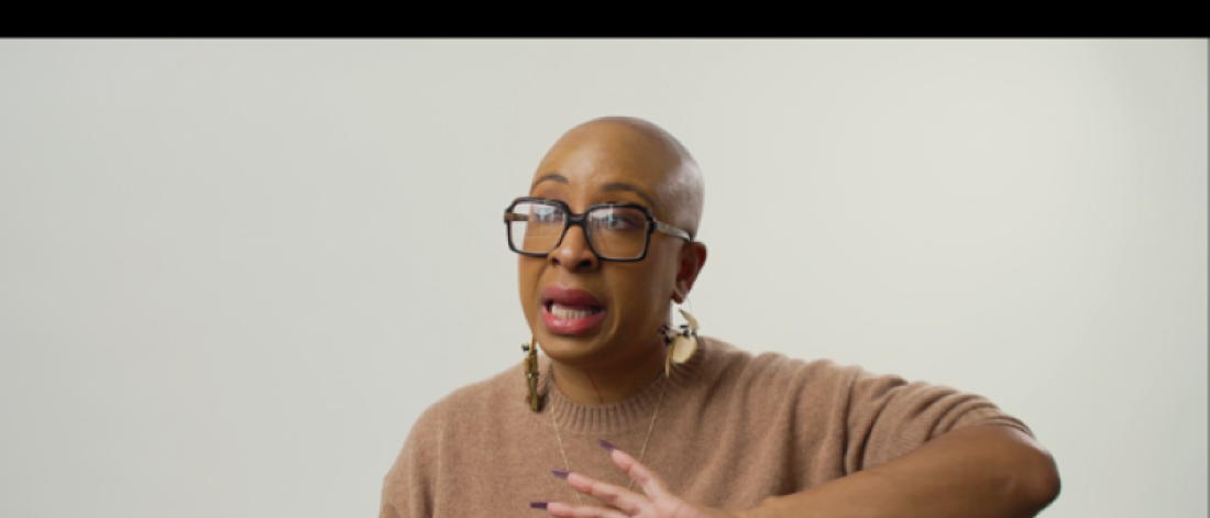 Negarra A. Kudumu wears a beige sweater, dark rimmed glasses, and chunky gold earrings. She is sitting down facing the camera against a neutral wall, and her hand is gesturing in front of her.