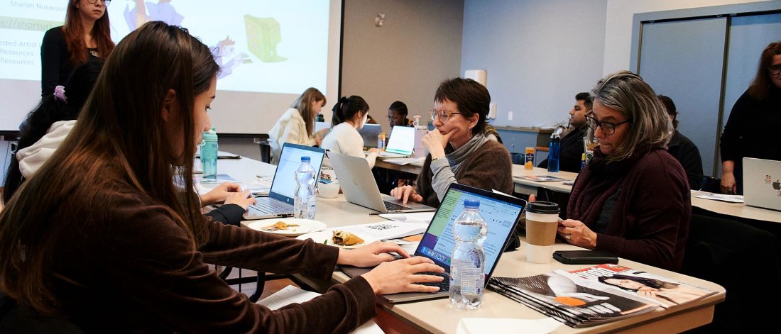 A group of people work on laptops around rectangular beige tables in a room. A person stands at the front in front of a projected image on a screen.