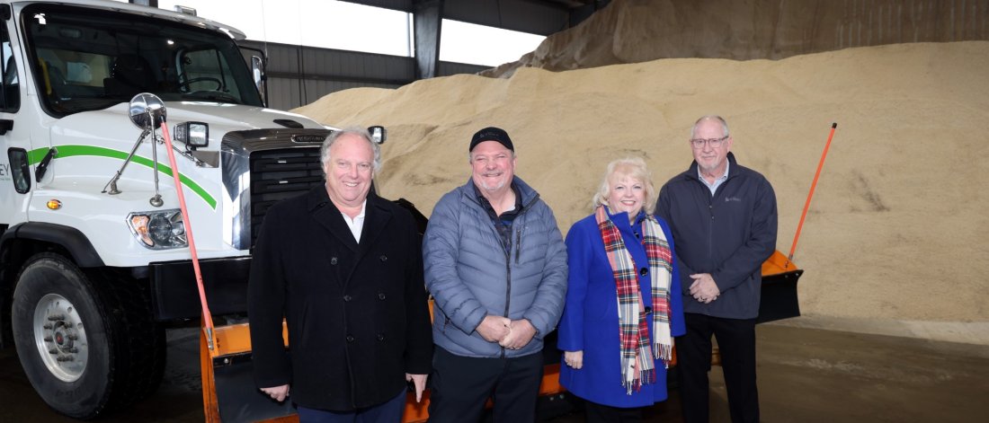 Four people posing in front of a snowplow truck inside a facility with a large pile of sand or salt in the background.