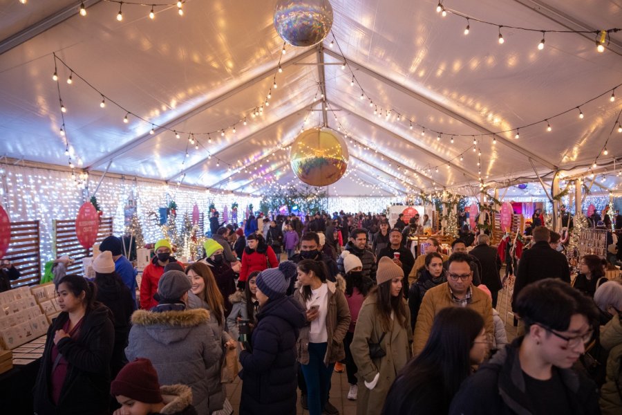  A busy holiday market under a tent decorated with string lights and festive decorations, with shoppers browsing through the stalls.