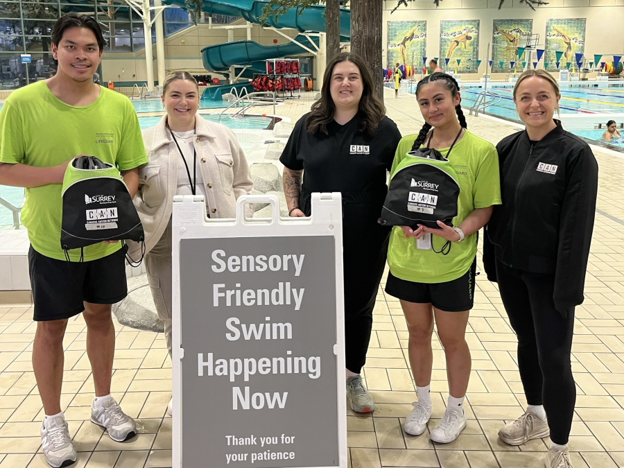 Staff smiling next to a sign that says, "Sensory friendly swim happening now," in a pool.