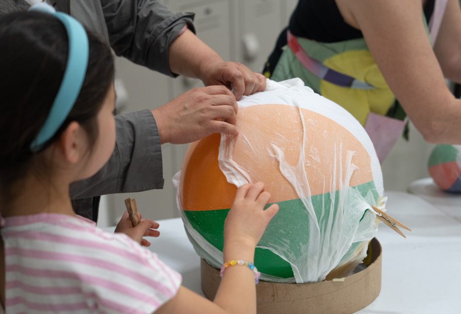 A young girl with a headband assists an adult in crafting a large, multicolored papier-mâché globe at a workshop table