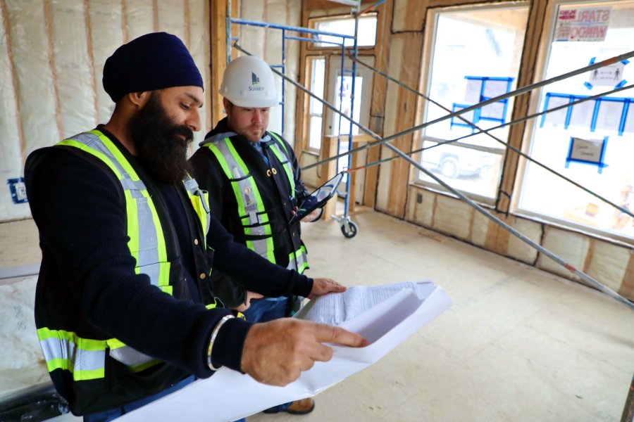 Two construction supervisors, one wearing a turban, reviewing blueprints inside a building under construction, illuminated by natural light from large windows.