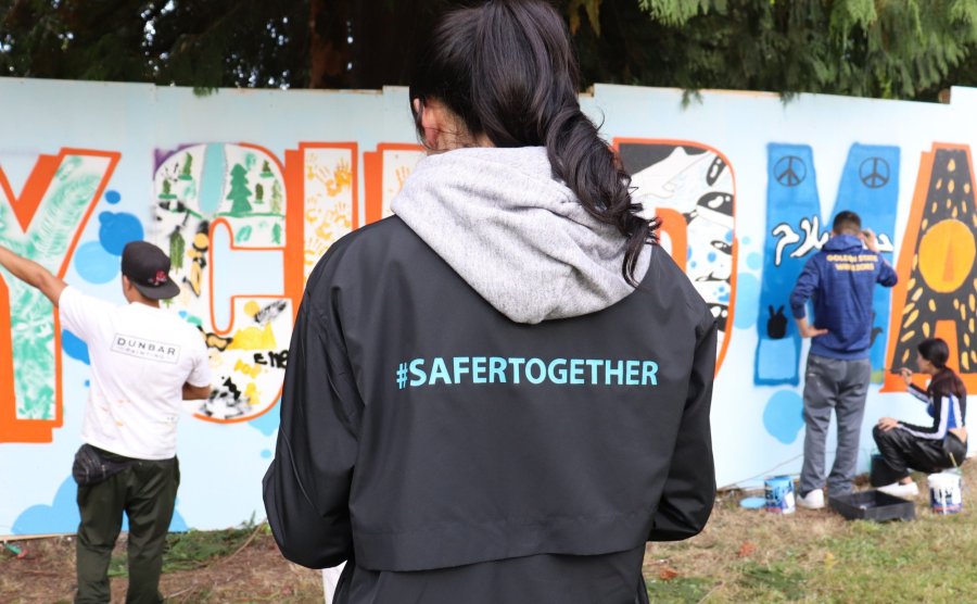 A person in a jacket with "#SAFERTOGETHER" printed on the back observes a mural being painted by community members.