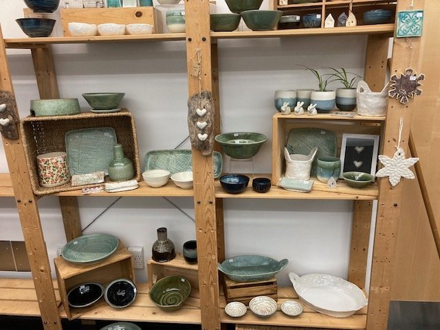 Assorted handmade ceramic pottery on wooden shelves, including bowls, plates, and decorative items in various styles and colors.