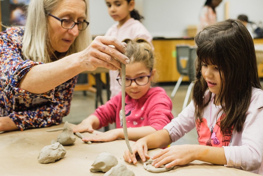 An instructor holds up a long coil of clay in front of two students working with clay at a table.