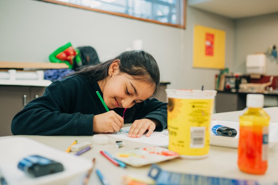 A child leans over a desk with art supplies on it, holding a green pencil and colouring on a paper.