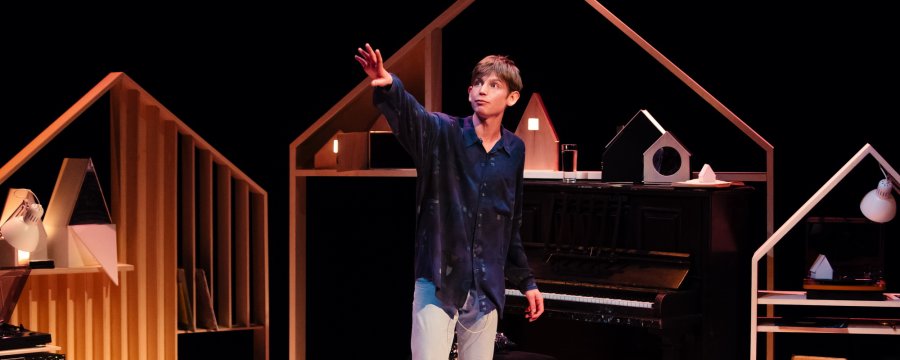 Performer Toby Thompson on stage arm raised in front of a set that includes a piano and wooden pieces that resemble the inside of a house 