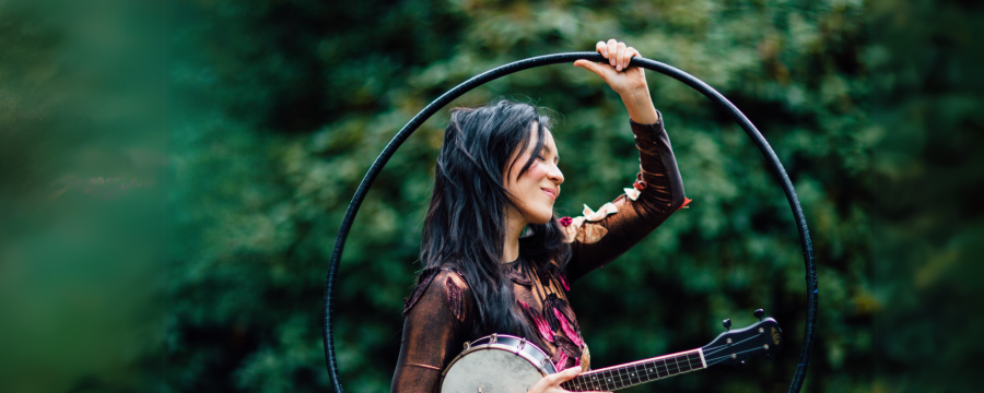 Performer Ginalina looks off camera as she stands in front of greenery while she holds her banjo and a hula hoop