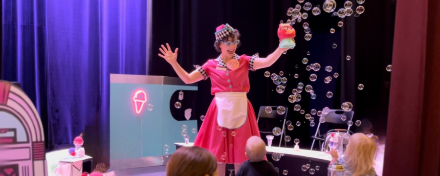 Actor dressed in a 1950s diner style waitress costume stands on the stage in front of audience while she holds a bubble making machine with bubbles coming out of it.