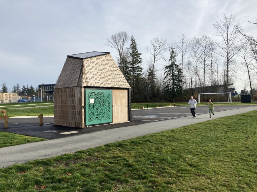 A small wooden structure with artistic designs, situated in a park with people jogging nearby.