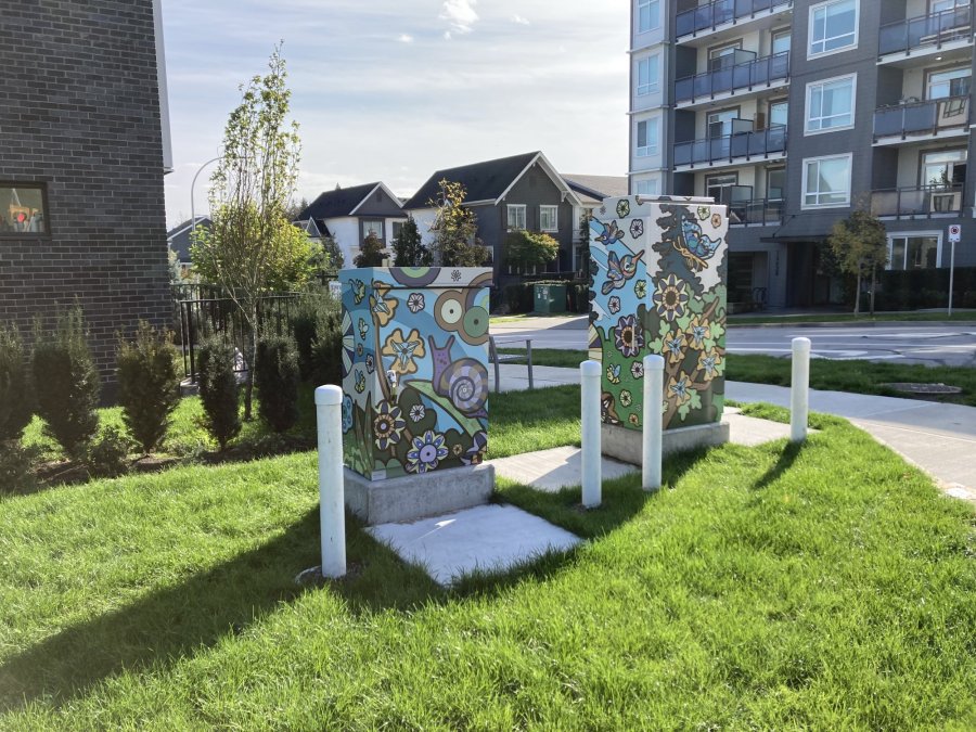 Decorated utility boxes featuring colorful floral and animal-themed artwork, situated in a green space near residential buildings.